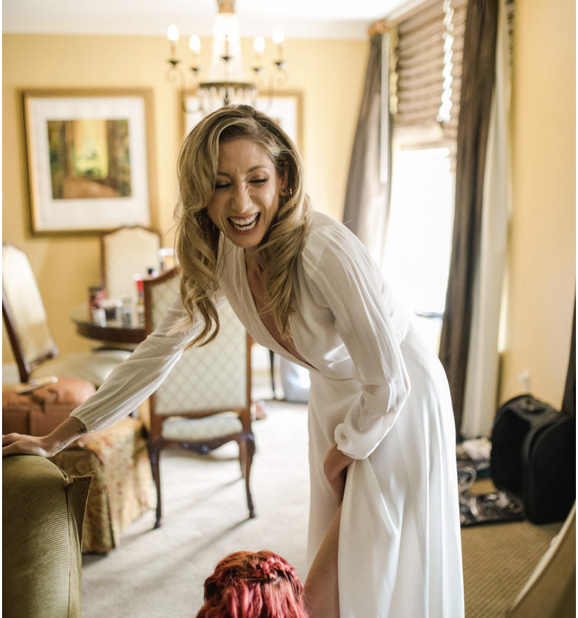 A laughing bride in a white dress leans down to adjust her shoe, assisted by a person with red hair.