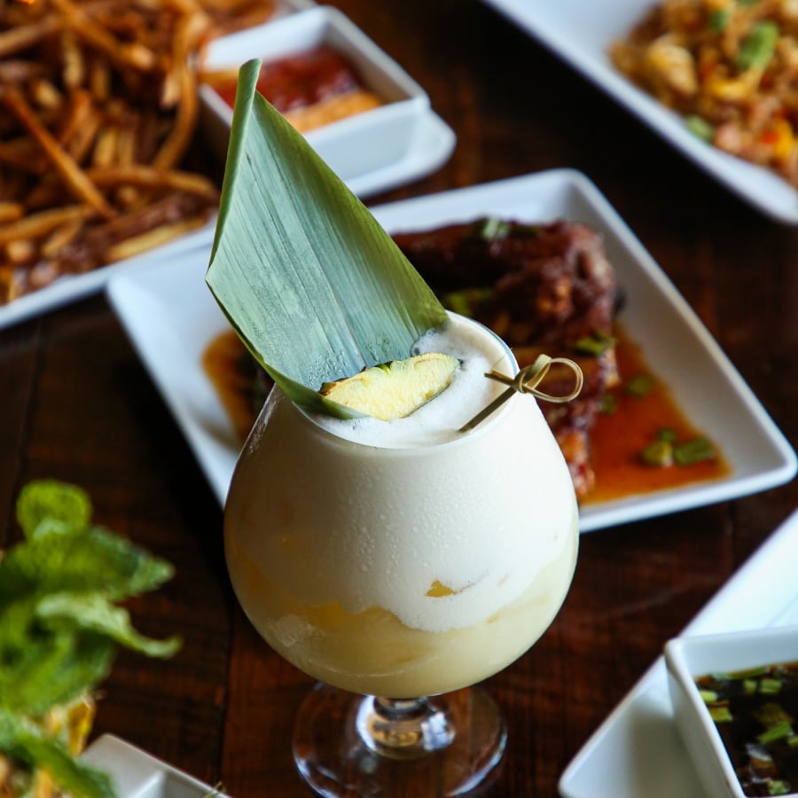 A creamy white tropical cocktail garnished with a banana leaf and lime, surrounded by appetizers and fried food.