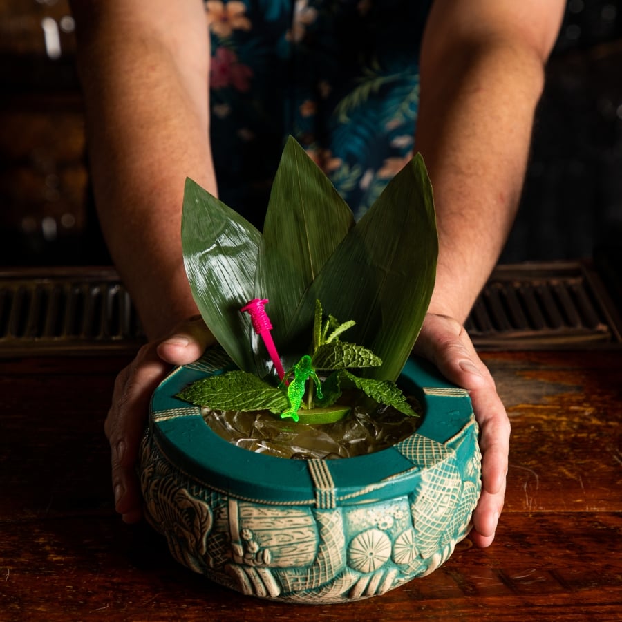 A bartender holds a large, teal tiki mug garnished with mint, leaves, and a tiny pink plastic toy.