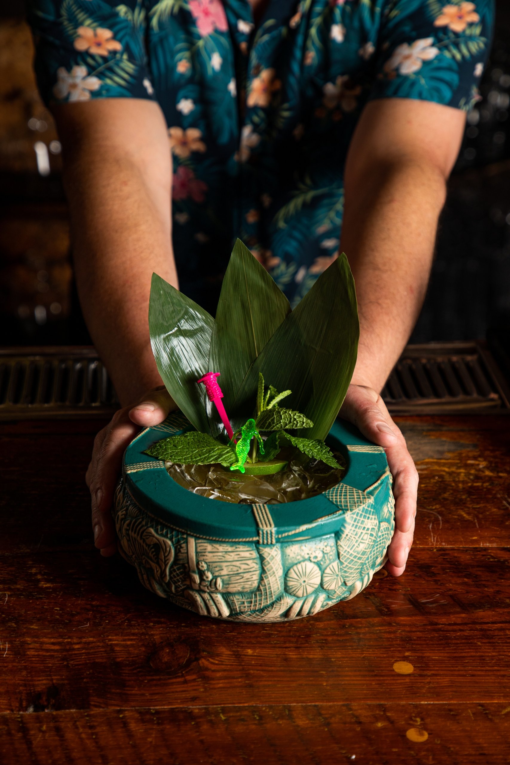 A bartender holds a large, teal tiki mug garnished with mint, leaves, and a tiny pink plastic toy.