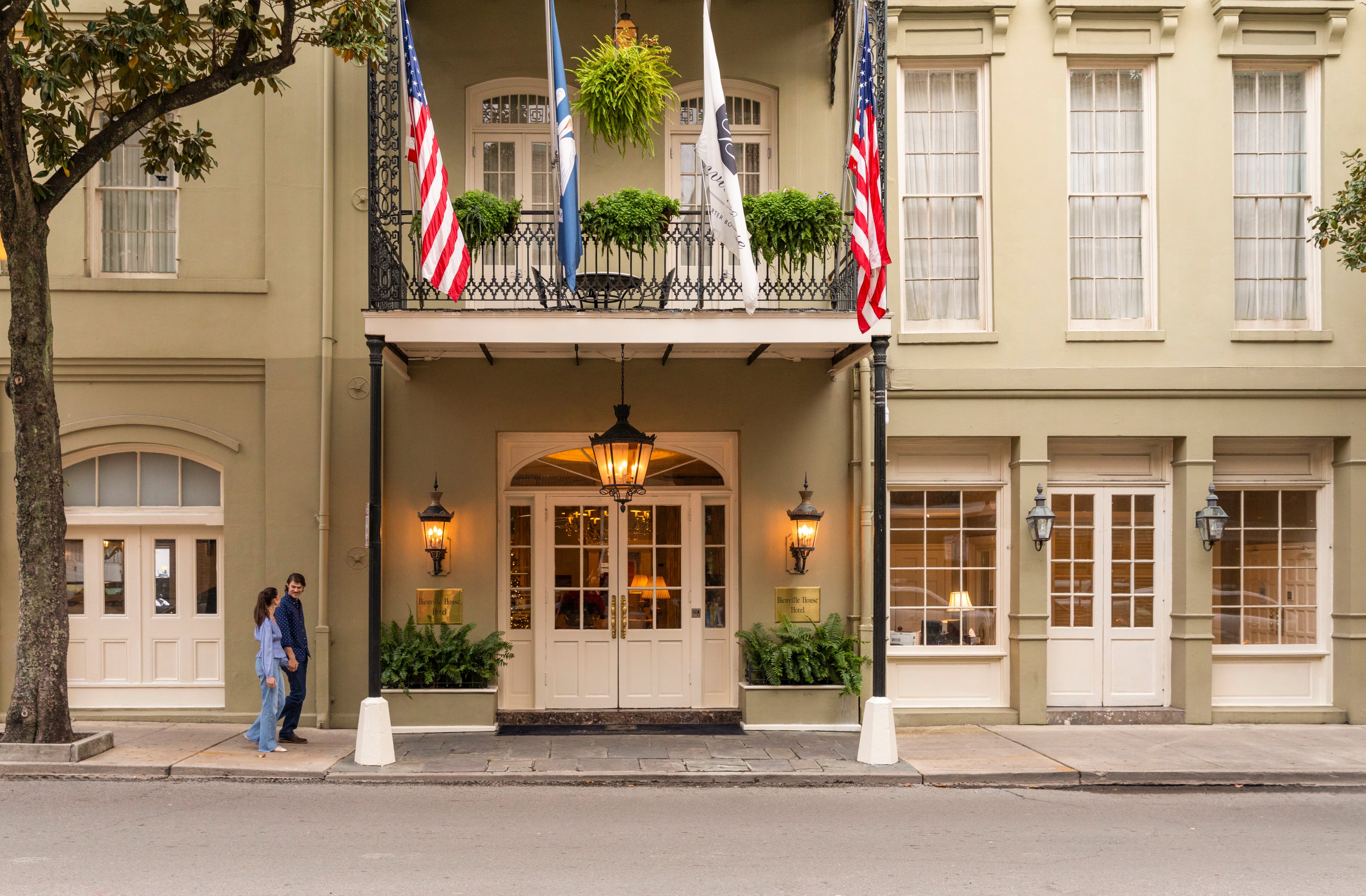 A beige building with a balcony, flags, and lights, with two people walking by.
