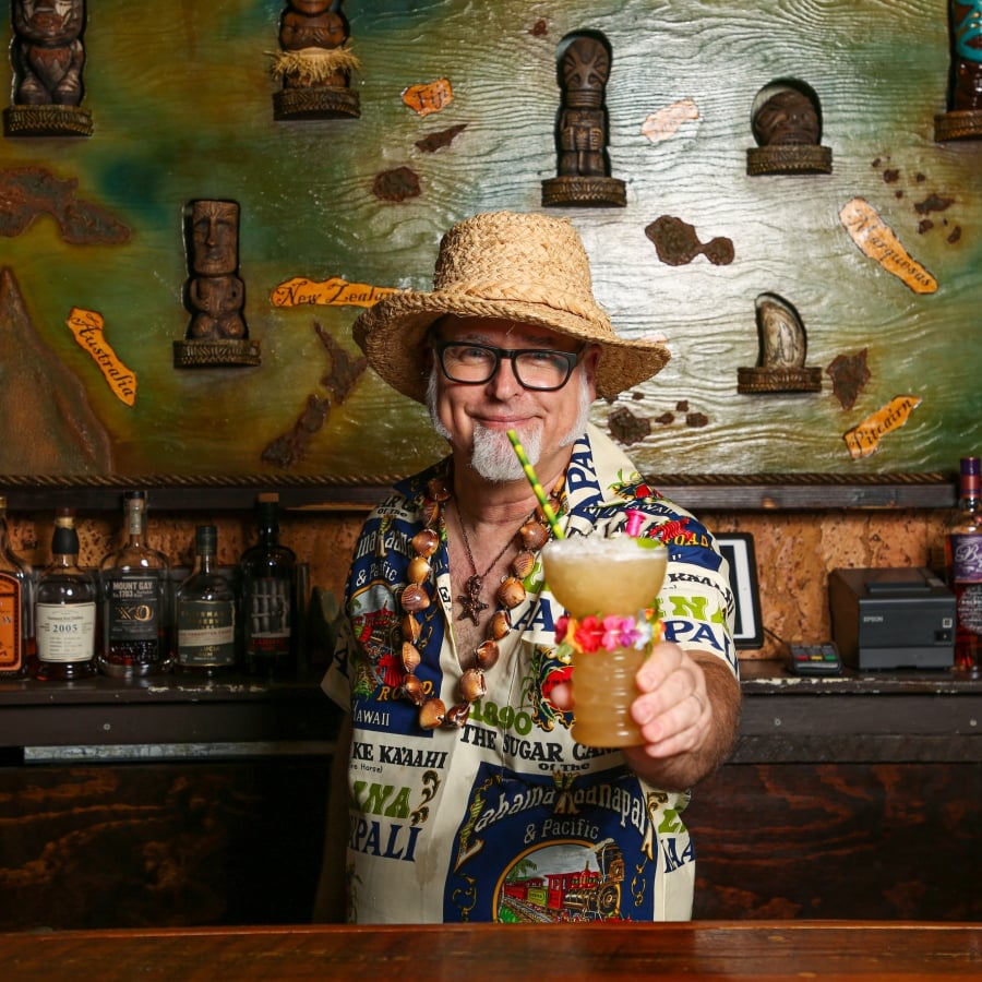 A bartender in a straw hat and Hawaiian shirt holds out a tiki drink, standing in front of a carved wooden map.