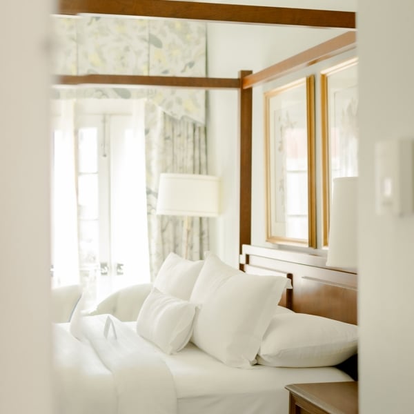 A bright, soft-focus view of a hotel room bed and carved wooden bedposts, seen from the hallway entrance.