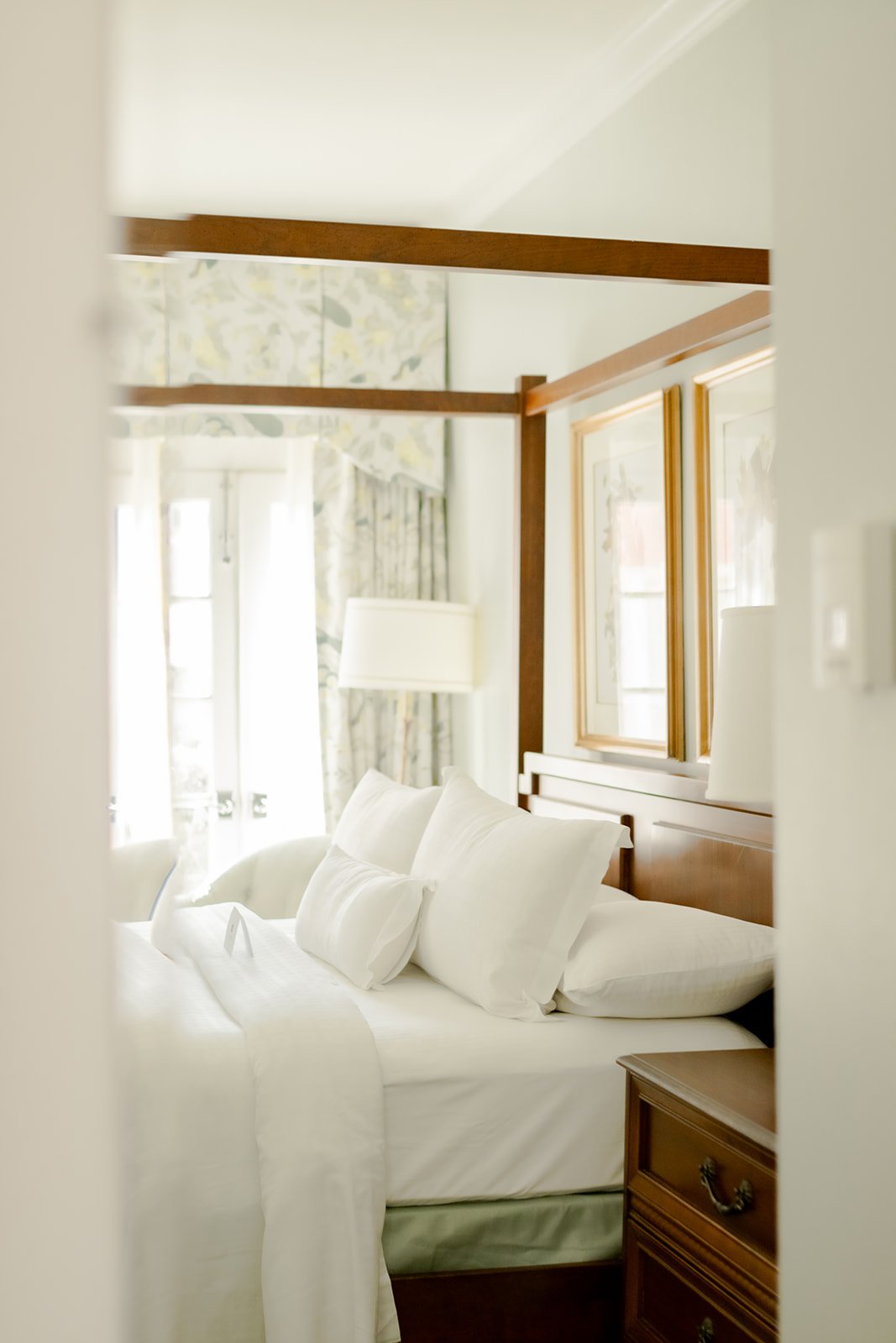 A bright, soft-focus view of a hotel room bed and carved wooden bedposts, seen from the hallway entrance.