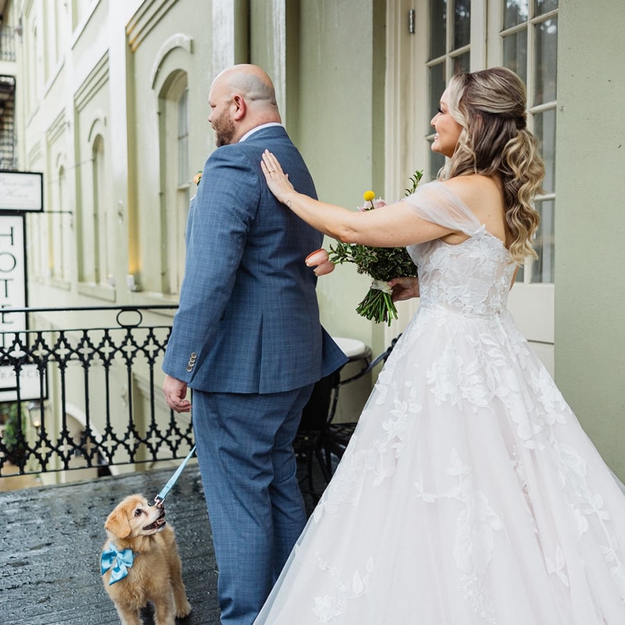 A bride gently taps her groom's shoulder while a golden retriever puppy stands nearby on a leash.