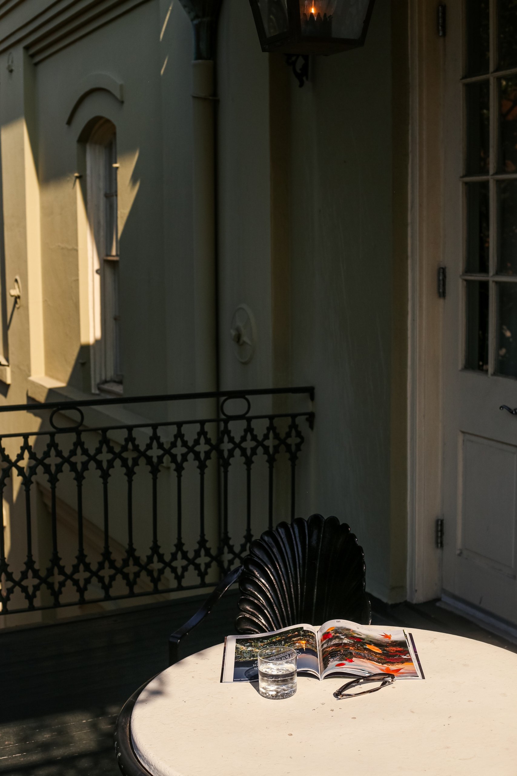 A small outdoor table on a balcony with a glass of water, an open magazine, and a black shell-back chair.