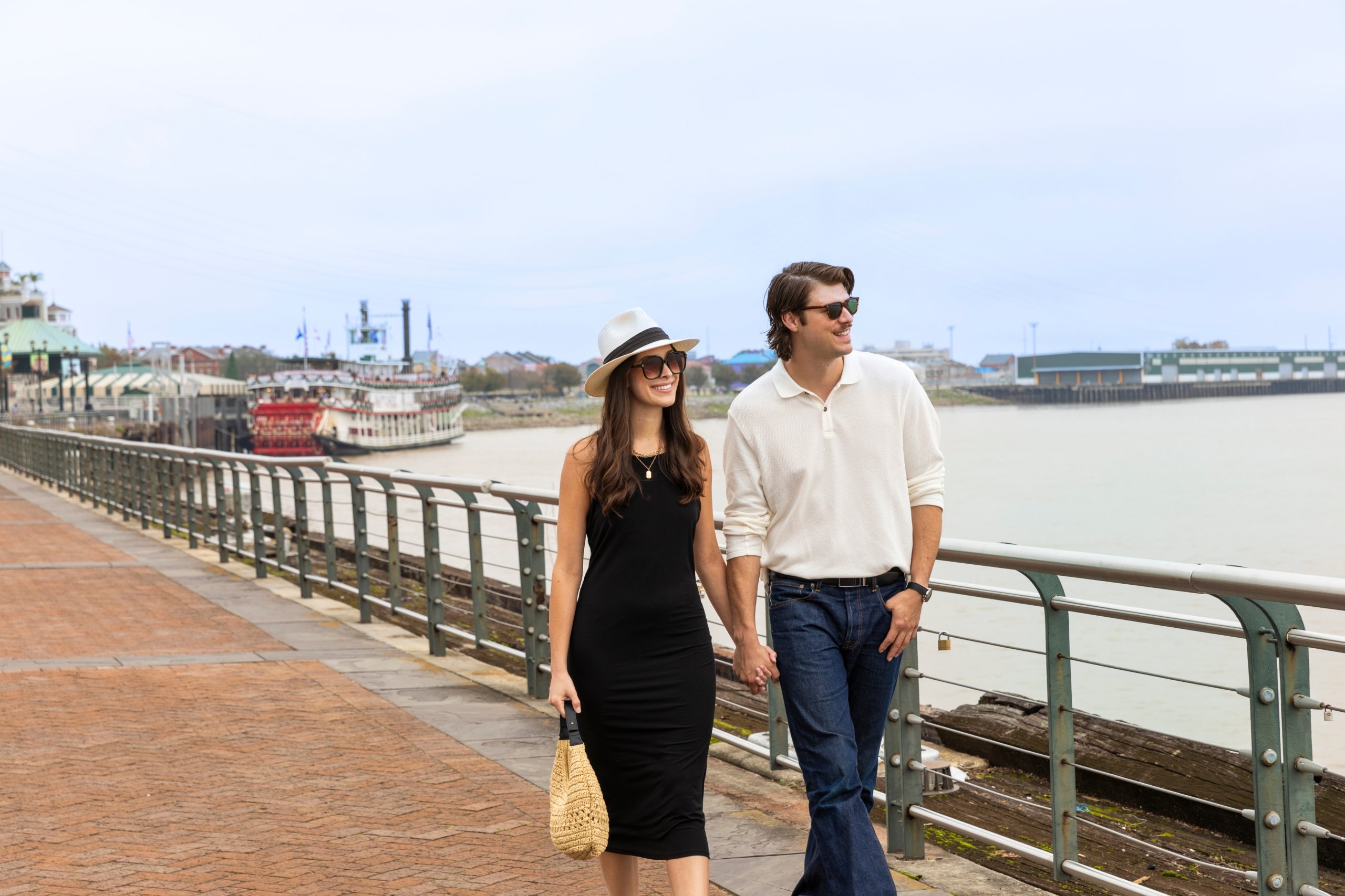 A couple holding hands walks along a brick promenade next to the Mississippi River with a steamboat visible.