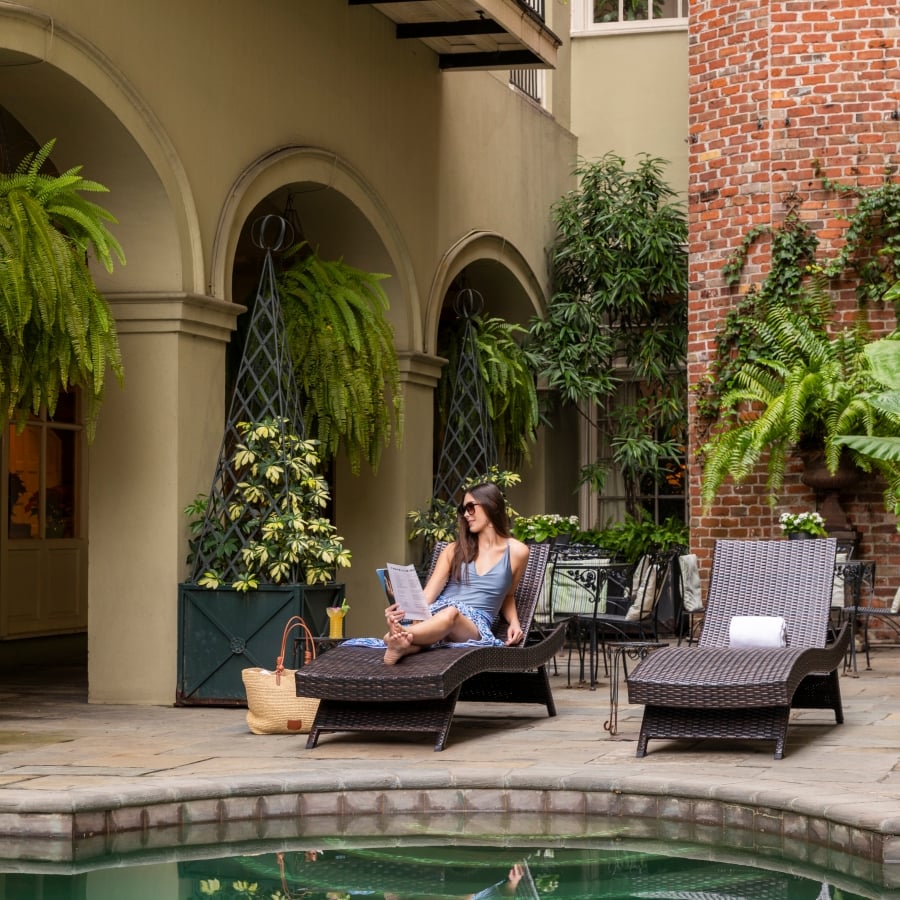 A woman in sunglasses reads a menu while relaxing on a woven chaise lounge by the courtyard pool.