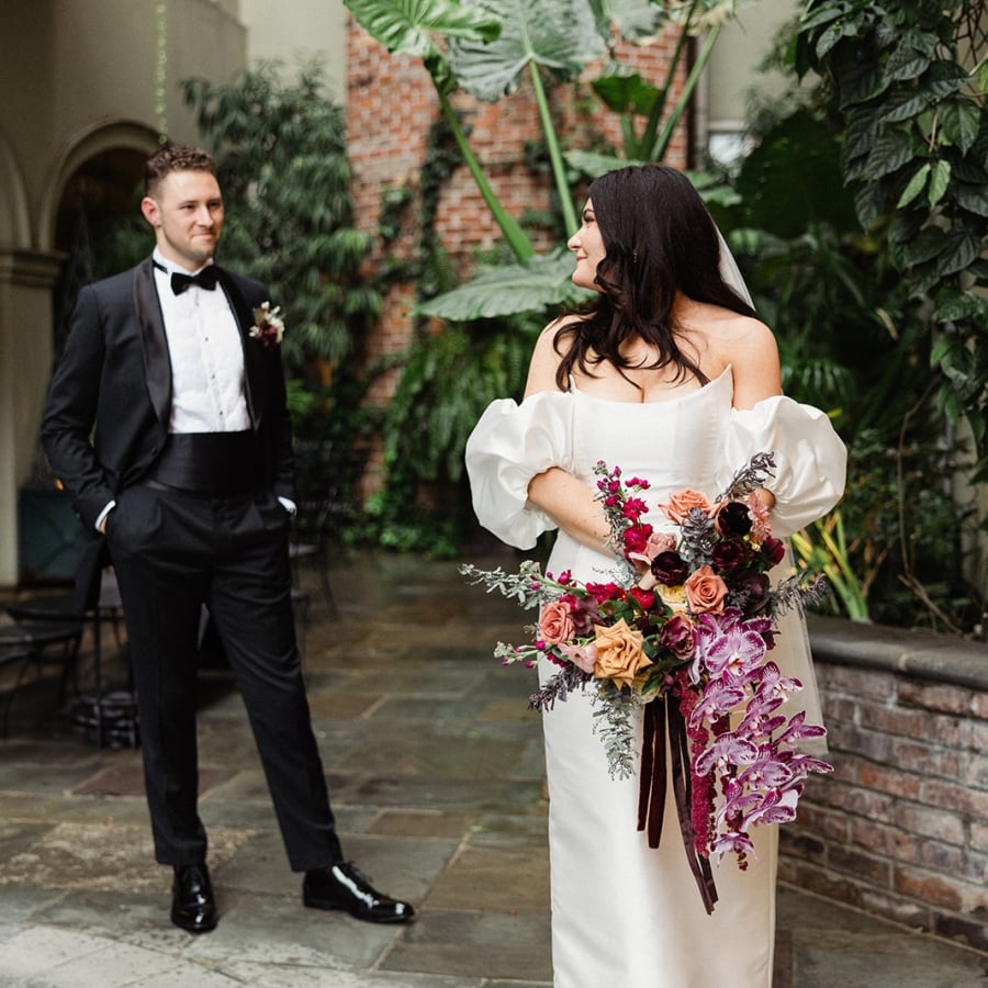 A bride in an off-shoulder gown with a dramatic bouquet poses by a pool as the groom stands in a tuxedo.