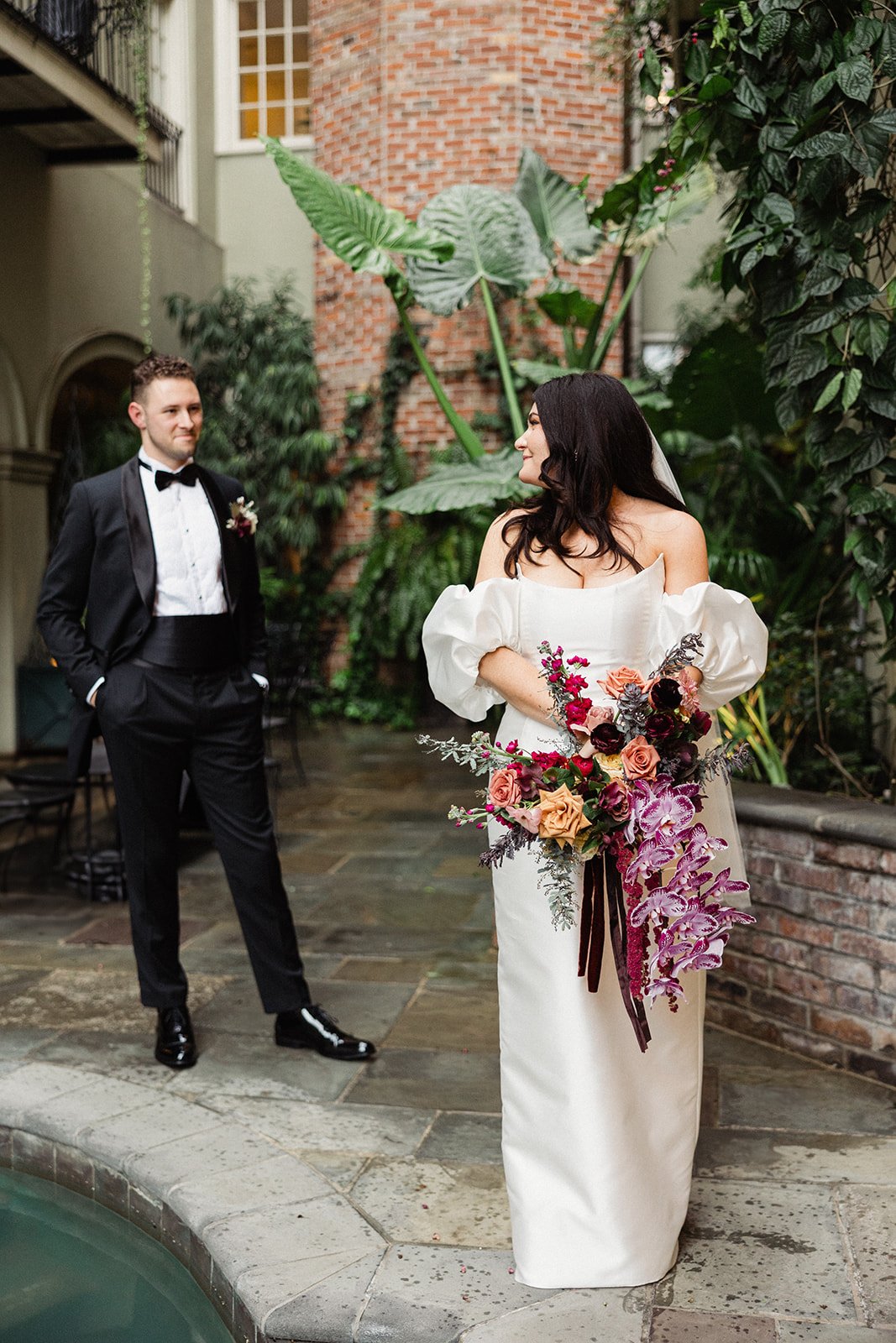 A bride in an off-shoulder gown with a dramatic bouquet poses by a pool as the groom stands in a tuxedo.
