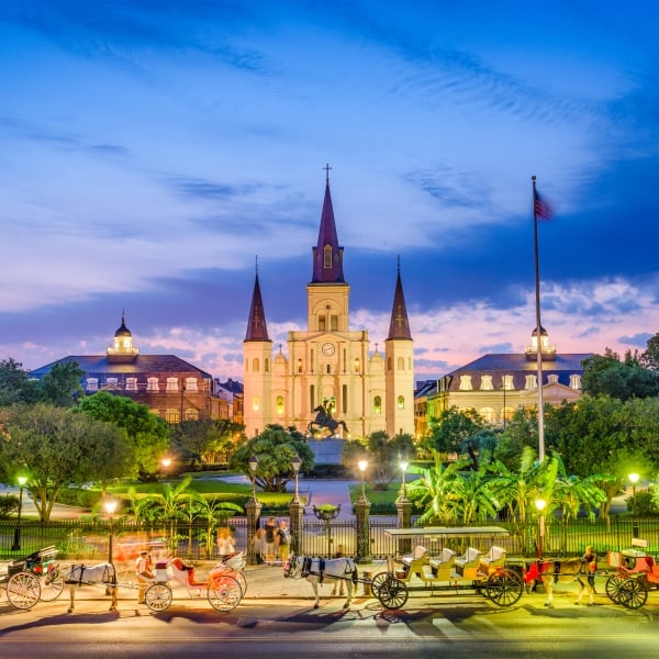 Jackson Square at dusk, featuring the illuminated St. Louis Cathedral and horse-drawn carriages in the foreground.