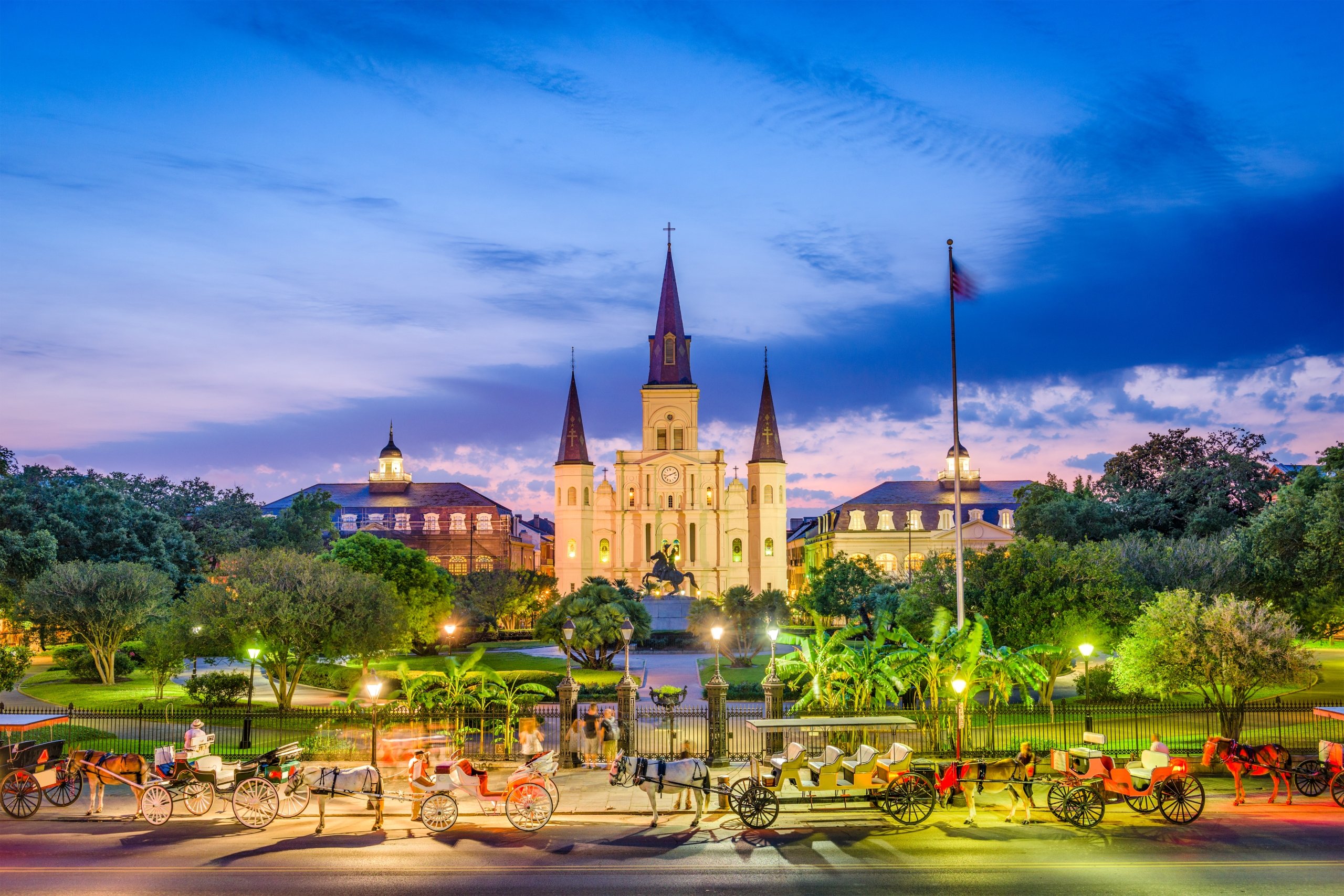 Jackson Square at dusk, featuring the illuminated St. Louis Cathedral and horse-drawn carriages in the foreground.