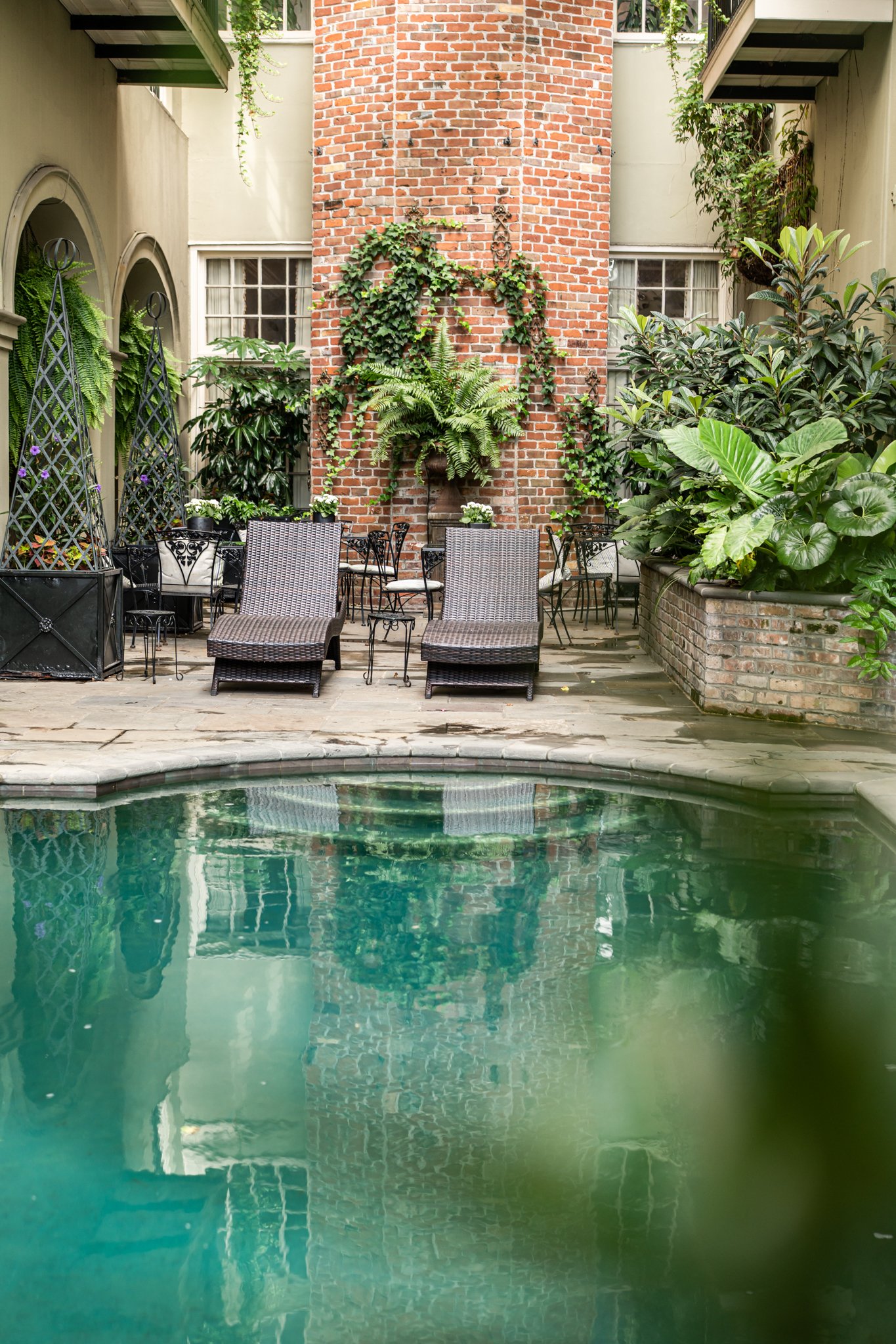Hotel courtyard with a reflective pool, brick chimney covered in ivy, and two woven lounge chairs.