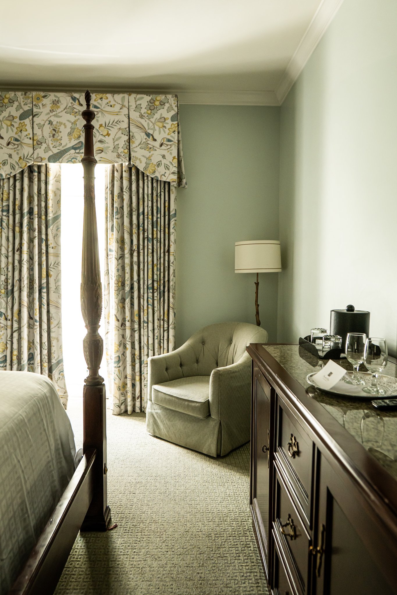 Hotel room corner with a wooden four-poster bed, patterned curtains, a mint-green wall, and an armchair.