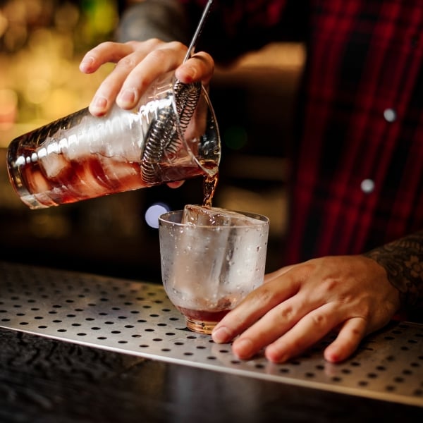 Bartender pouring a Vieux Carre cocktail from the measuring cup to a glass on the bar counter