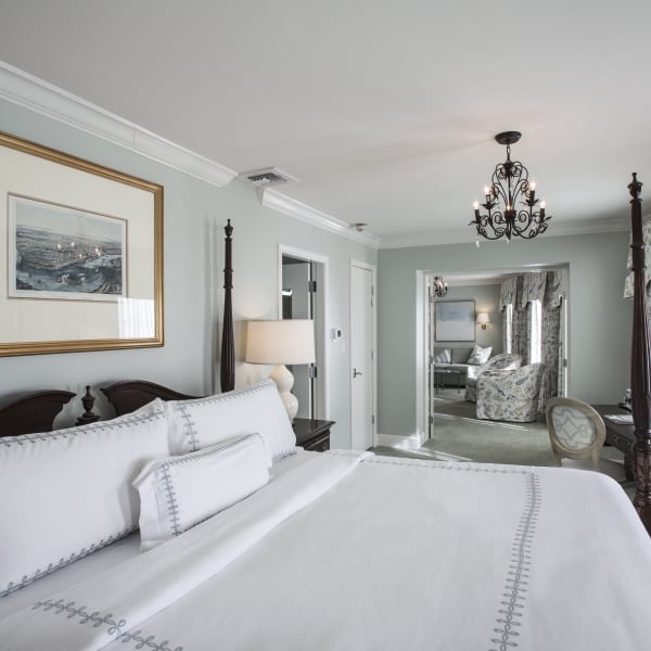 Bedroom with dark wood four-poster bed, white bedding with light blue embroidery, and a gold-framed cityscape artwork above. View extends to a sitting room.