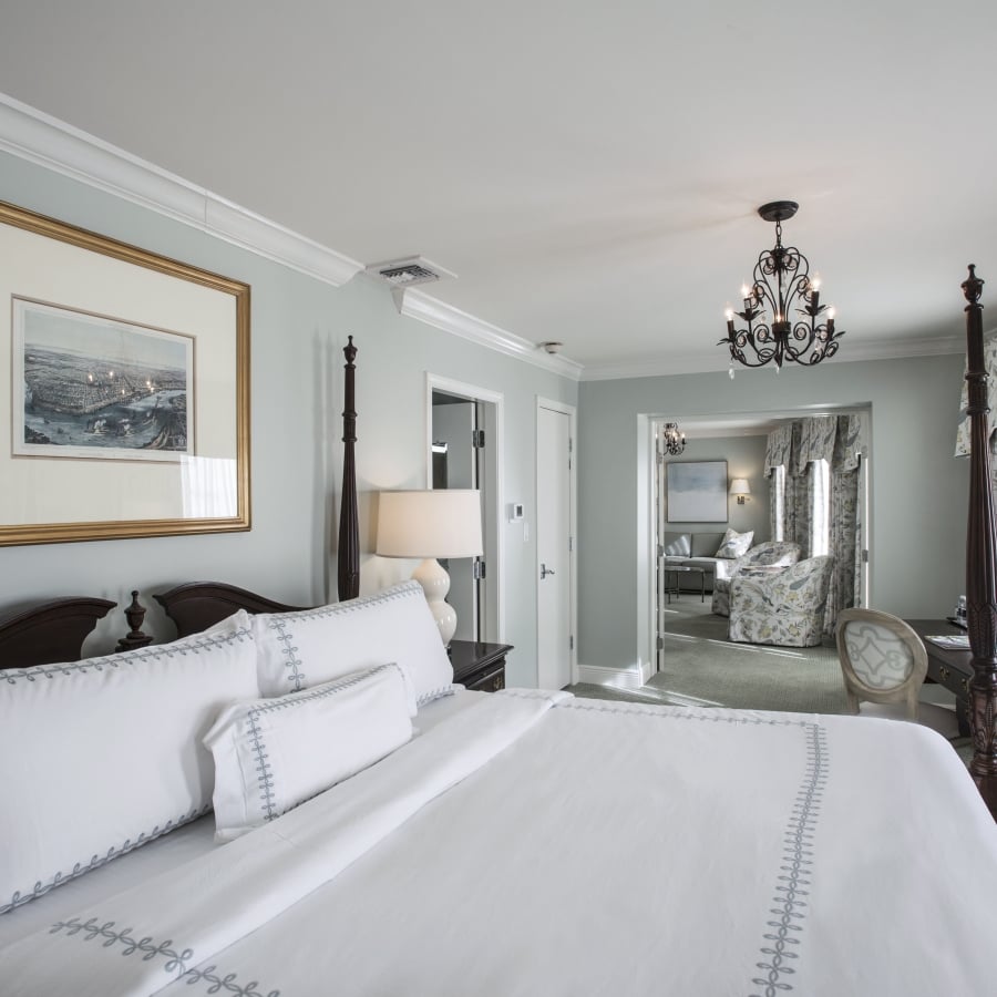 Bedroom with dark wood four-poster bed, white bedding with light blue embroidery, and a gold-framed cityscape artwork above. View extends to a sitting room.