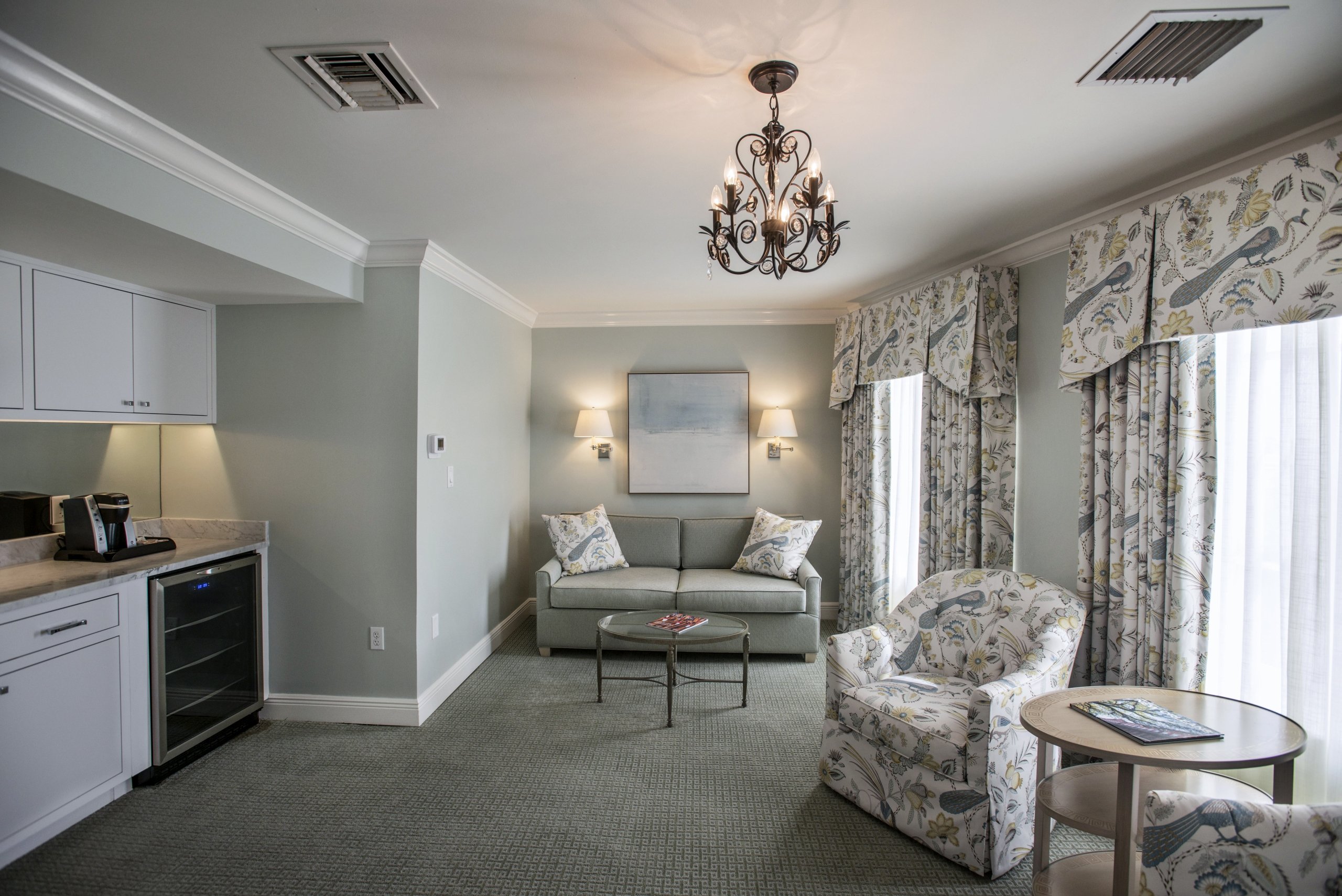 Suite living space showing a kitchenette with white cabinets and a beverage fridge, next to the sitting area with a sofa and armchairs.
