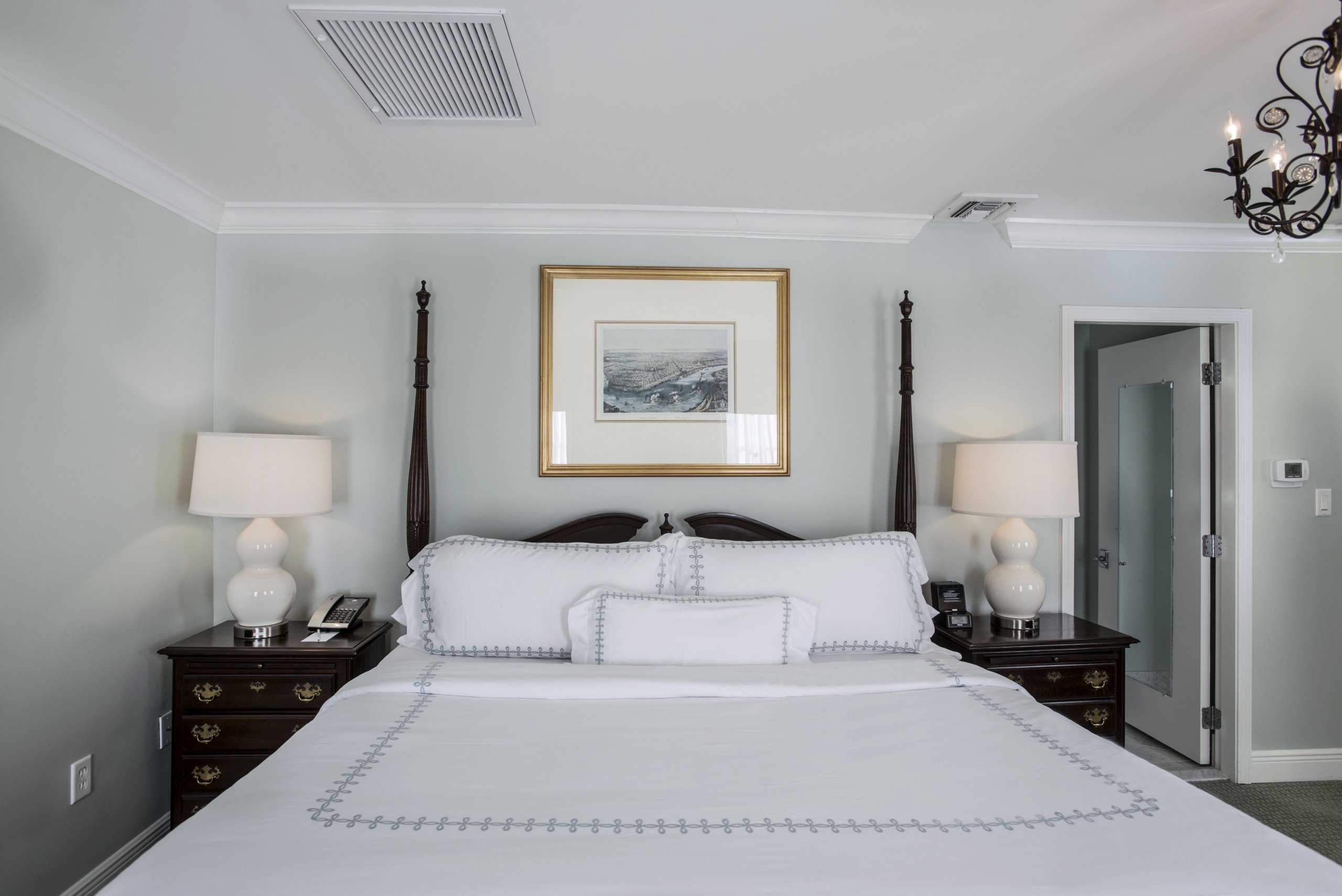 Head-on view of a king-sized dark wood four-poster bed with white linens, two dark wood nightstands with white lamps.