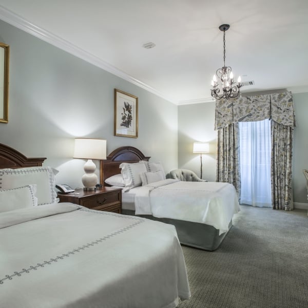 Deluxe hotel room with two wood-headboard double beds, white embroidered linens, patterned curtains, a desk, and bird-themed artwork.