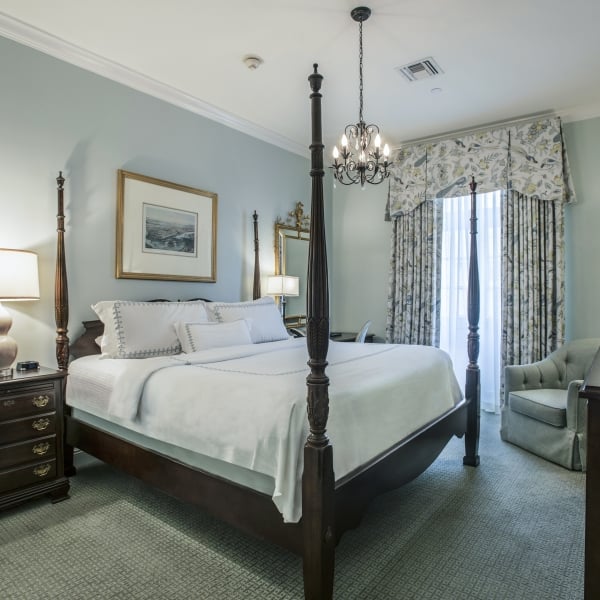 Deluxe King hotel room with a dark wood four-poster bed, light blue-green walls, patterned curtains, and a plush armchair by the window.