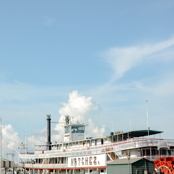 The white, multi-deck Steamboat NATCHEZ with its tall black smokestack and bright red paddlewheel, docked on the Mississippi River.