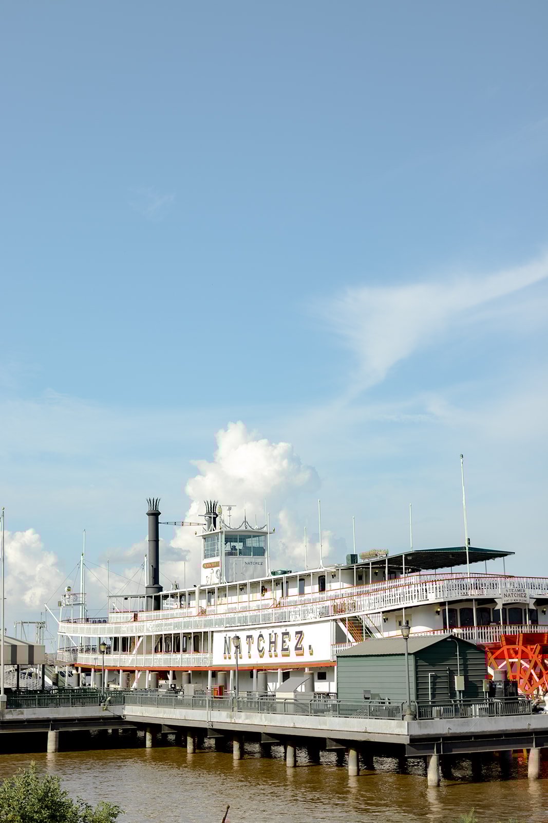 The white, multi-deck Steamboat NATCHEZ with its tall black smokestack and bright red paddlewheel, docked on the Mississippi River.