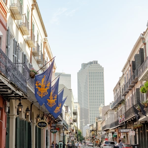 A street view in the French Quarter, showing buildings with wrought-iron balconies and fleur-de-lis flags, leading to modern high-rises.