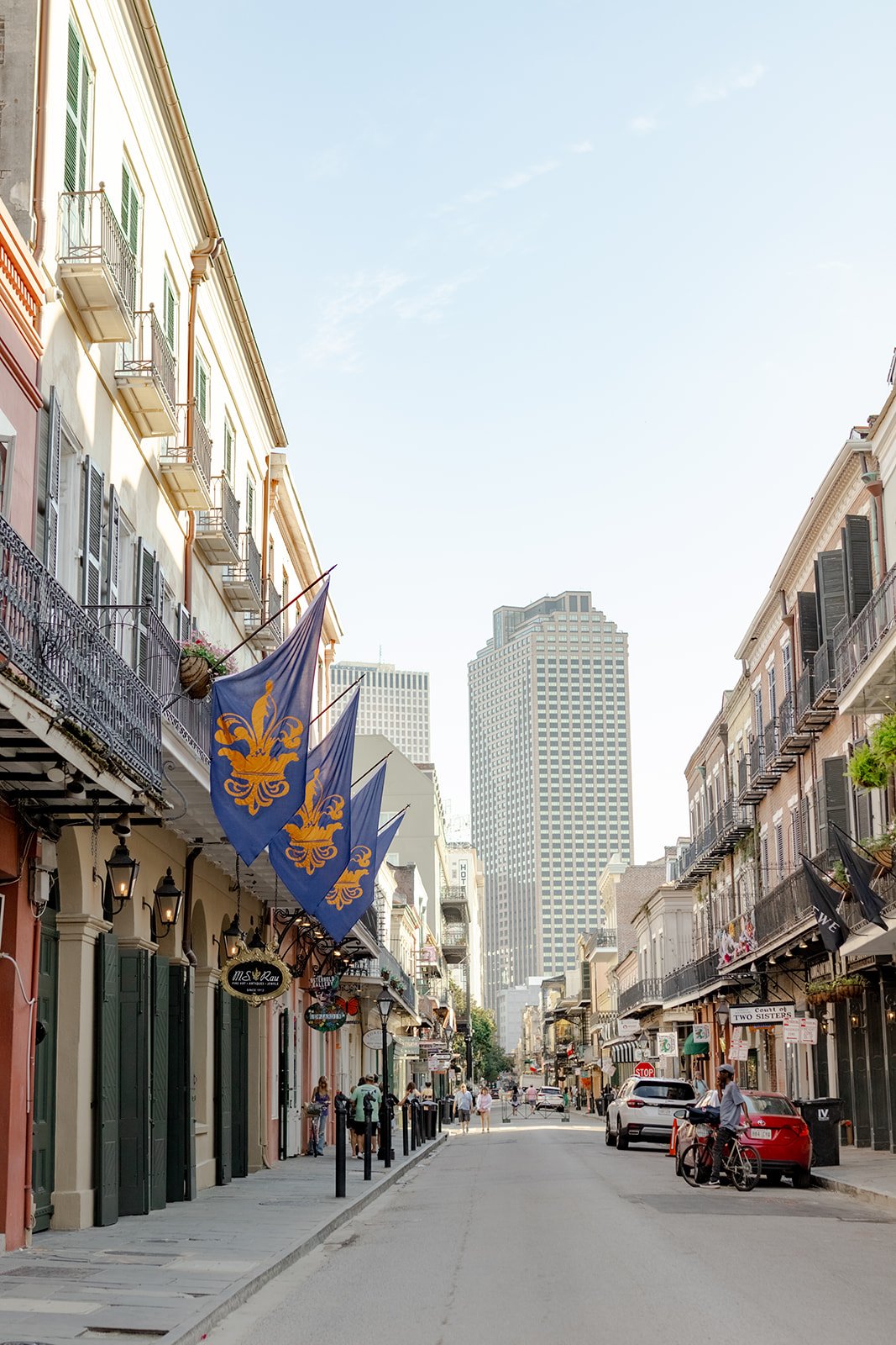 A street view in the French Quarter, showing buildings with wrought-iron balconies and fleur-de-lis flags, leading to modern high-rises.