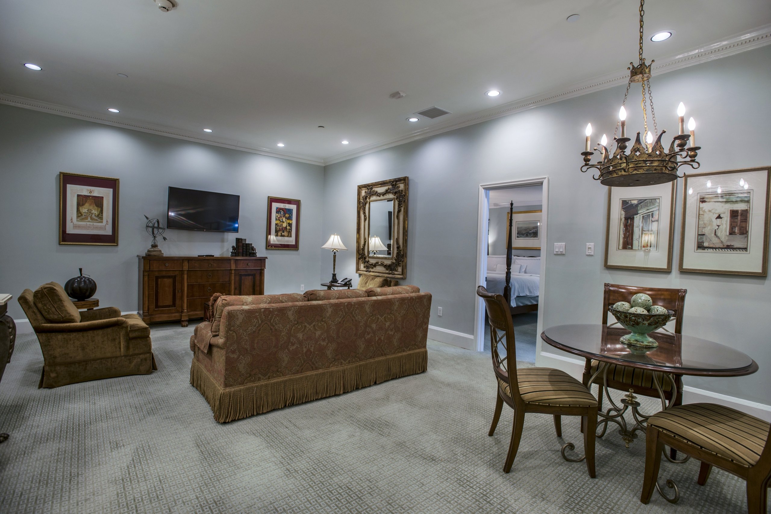A spacious hotel suite living room with a terracotta sofa, a dark wood dining table for three, and a wrought-iron chandelier.
