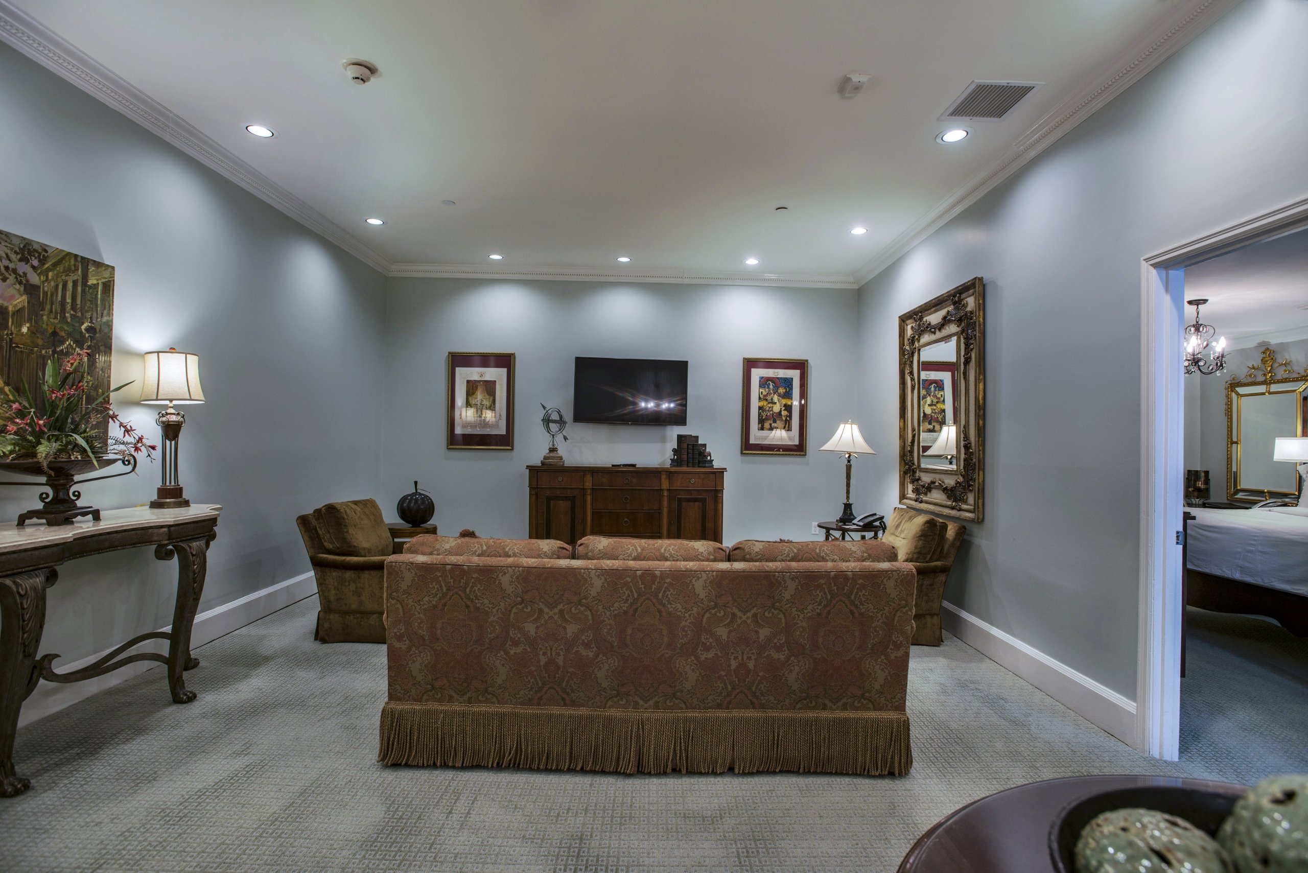 Living room area of a hotel suite featuring a terracotta sofa, a large flat-screen TV on a dark cabinet, and two upholstered armchairs.