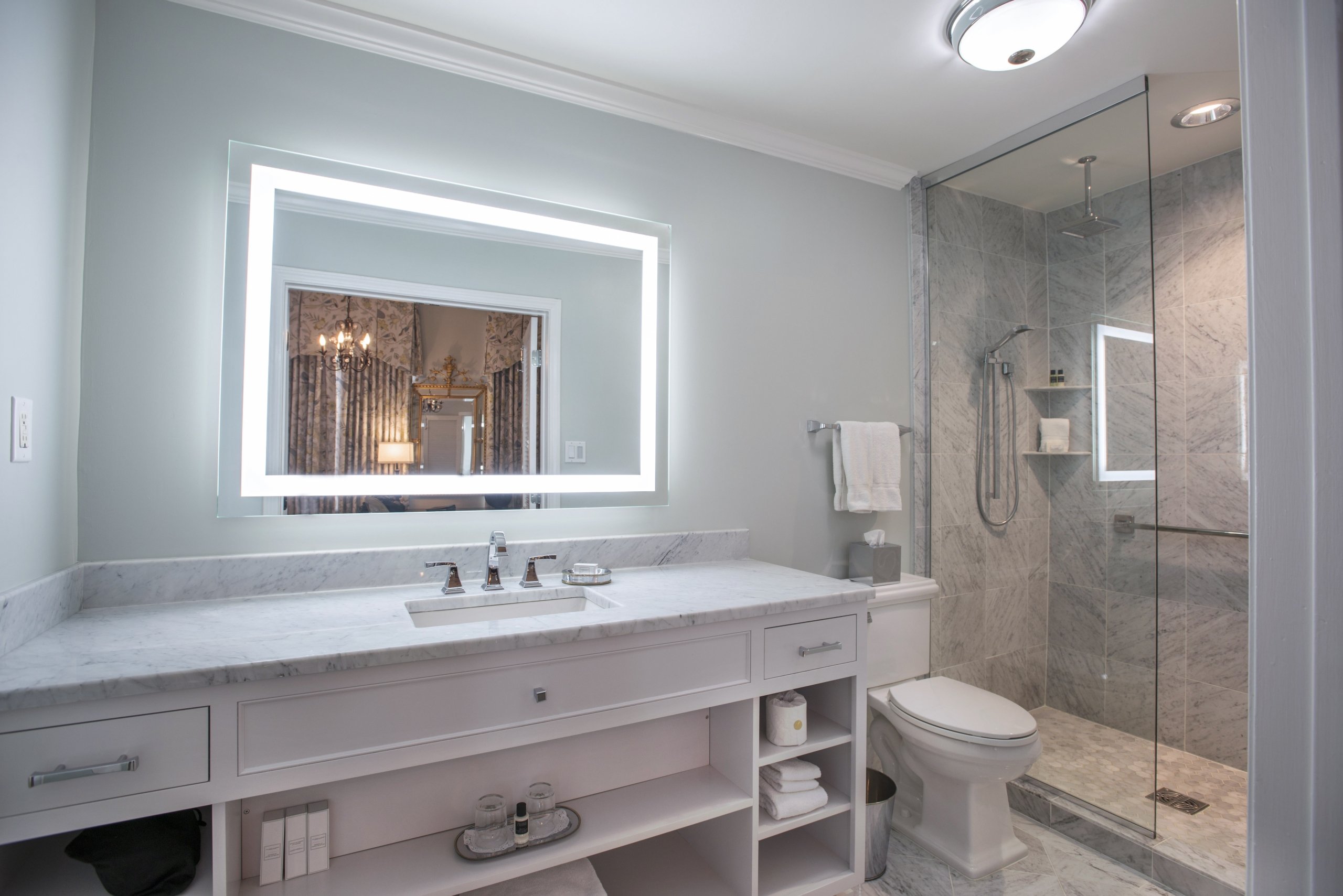 Renovated bathroom with a white marble vanity, an illuminated rectangular mirror, and a glass-enclosed shower with gray marble tiling.