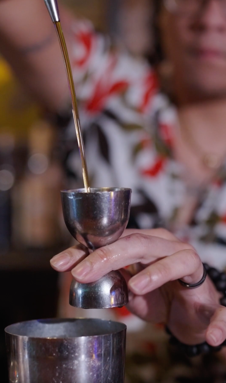 Close-up of a bartender pouring dark liquor into a metal jigger over a cocktail shaker.