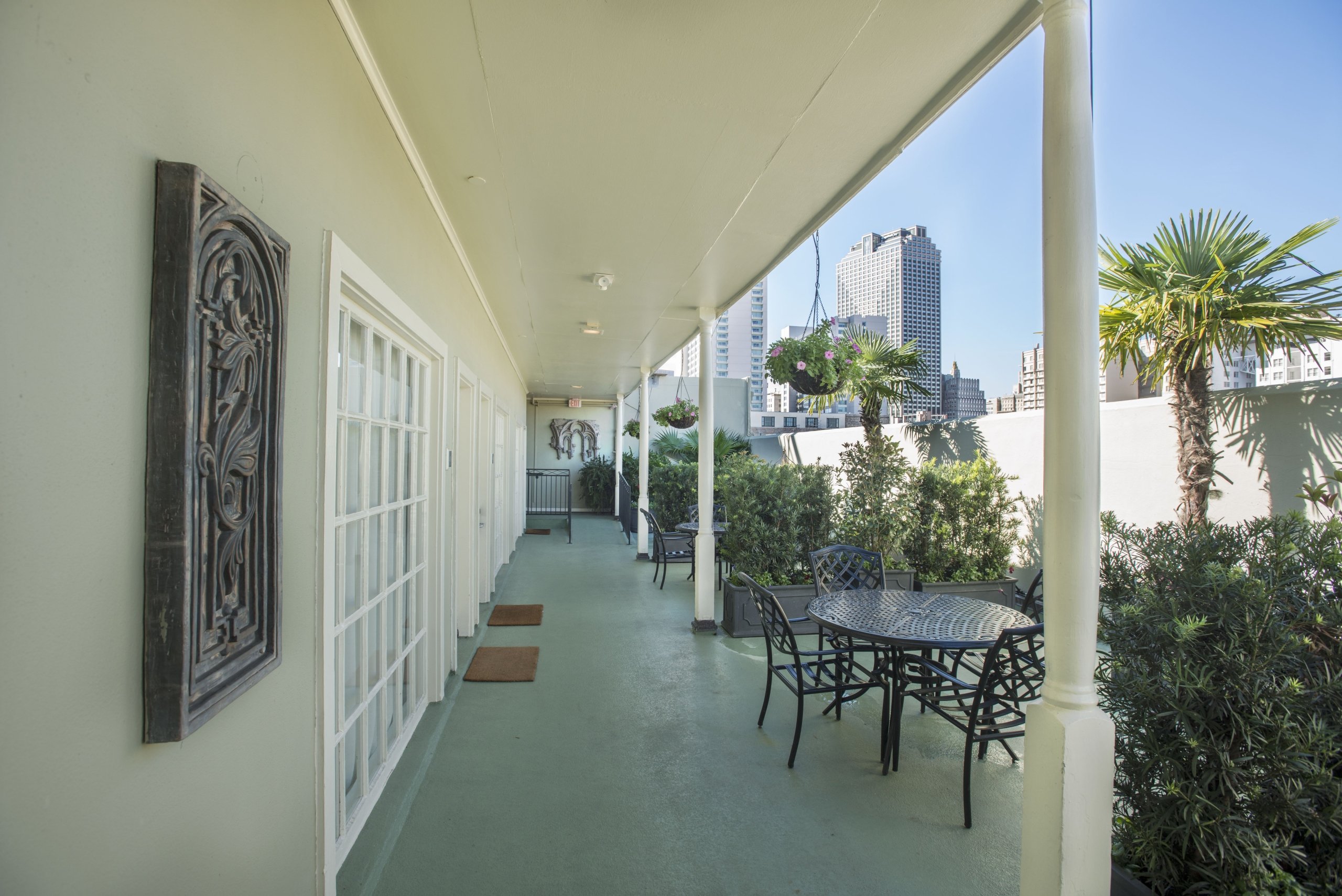 Long, covered outdoor sundeck patio with light green walls, metal tables and chairs, lush greenery, palm trees, and a view of city high-rises.