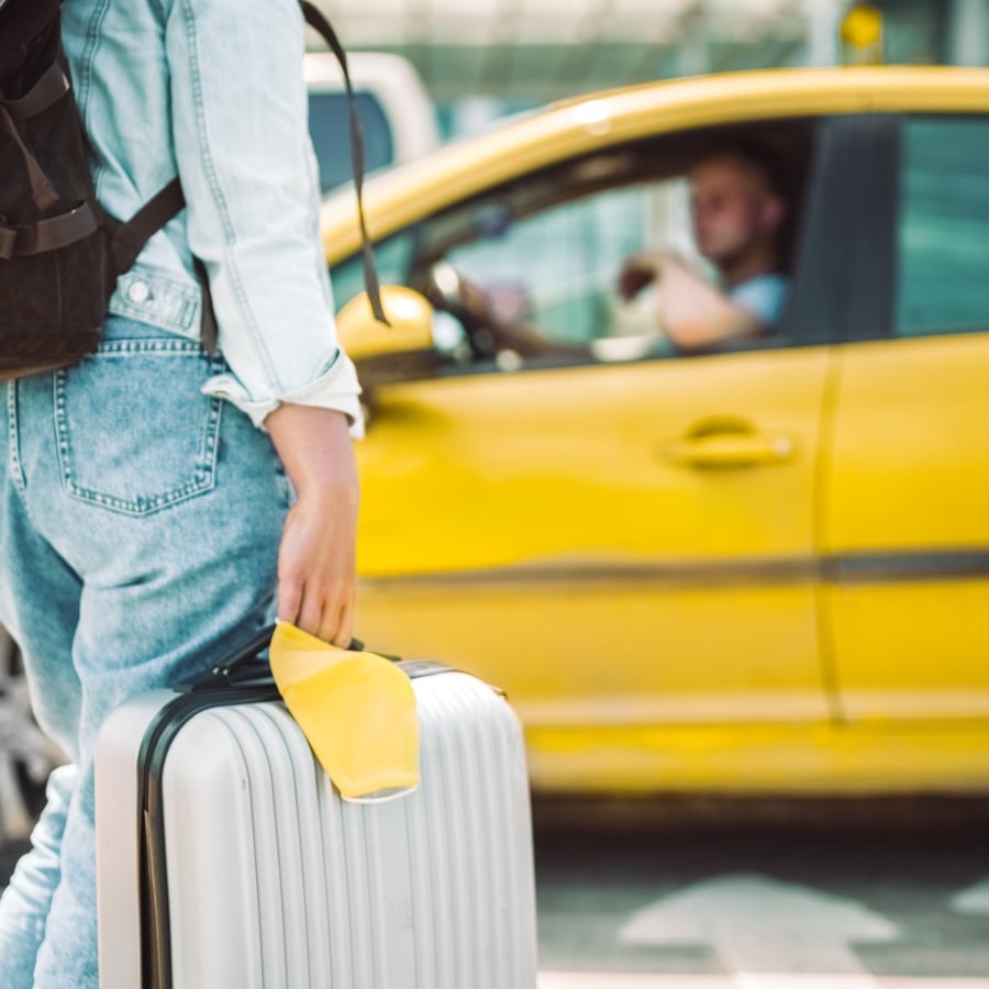 A person with a backpack and jeans walks away from a yellow taxi while pulling a rolling suitcase.