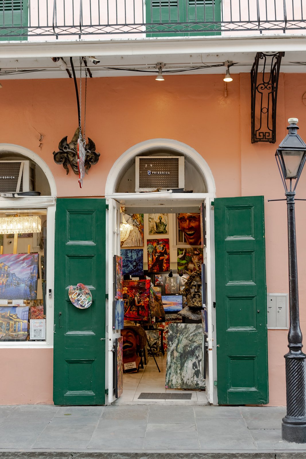 The arched entrance of a French Quarter art gallery with green shutters open to display colorful paintings inside.
