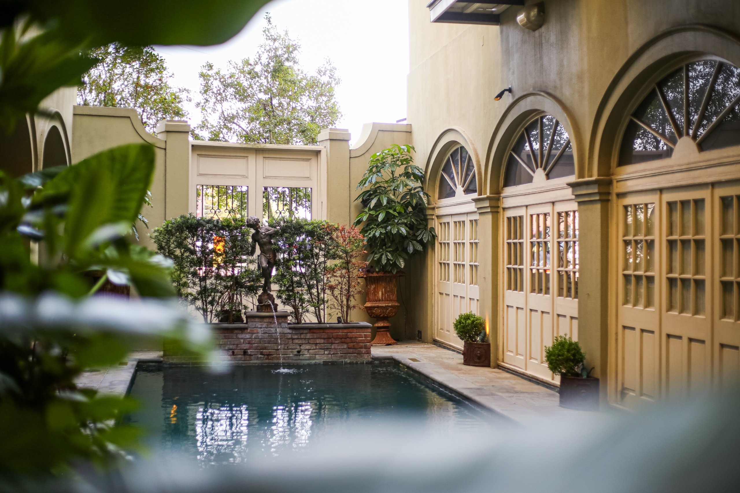 A secluded courtyard pool with a small fountain statue, arched openings, and lush plants, viewed through foreground foliage.
