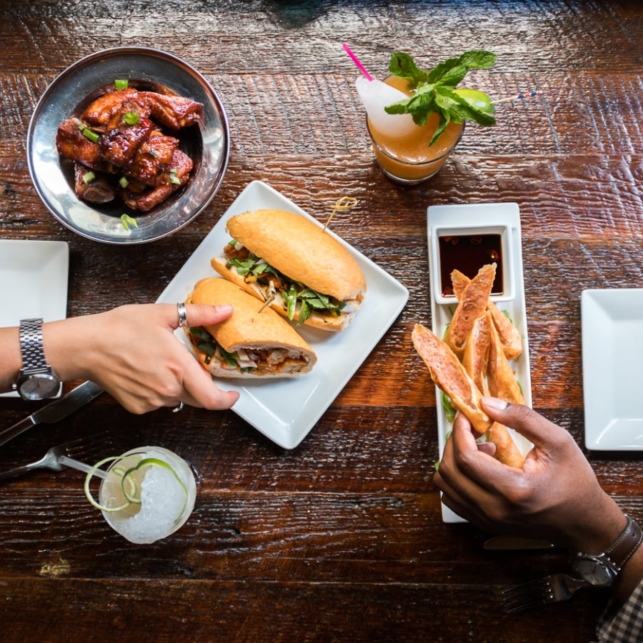 n overhead shot of hands sharing a Banh Mi sandwich and seasoned fries on a dark wooden table in a restaurant.