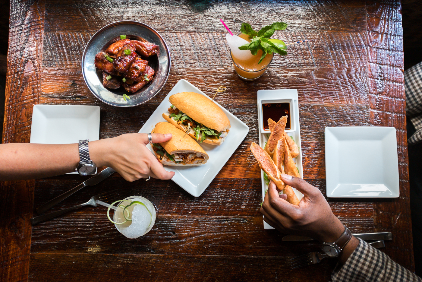 A table with food and drinks, with two people's hands reaching for sandwiches.