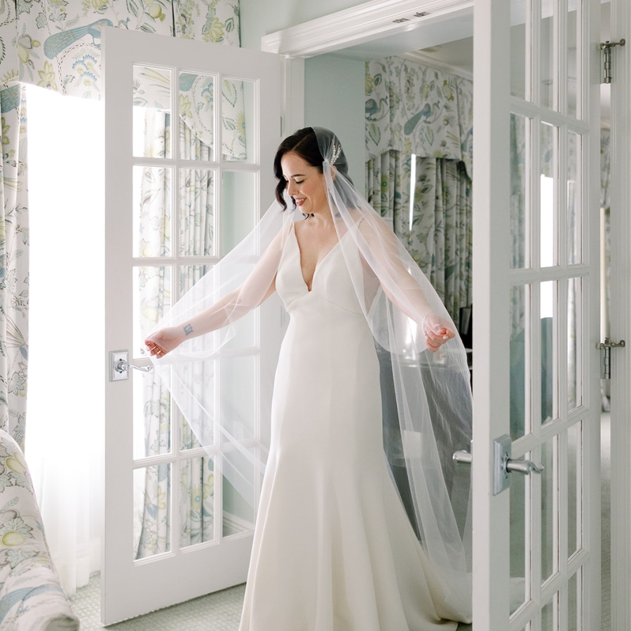 A bride in a fitted dress and veil stands between two white French doors opening into a bright hotel room.