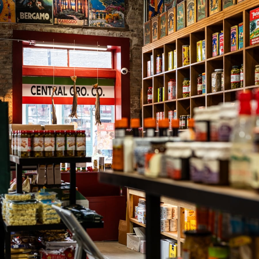 The crowded interior of an old-fashioned grocery store with shelves stocked with preserved goods and travel posters on the walls.