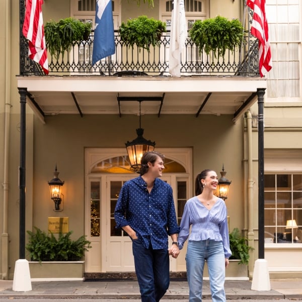 A smiling couple holding hands walks in front of the ornate entrance and balcony of the Bienville House Hotel.
