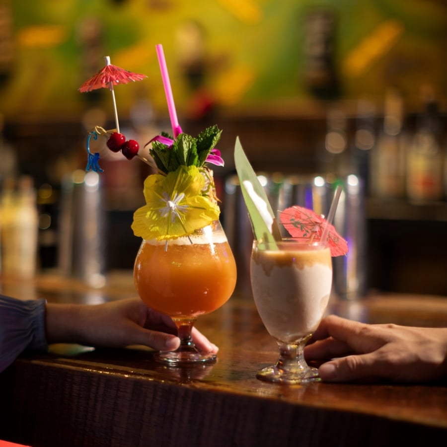 Two hands hold ornately garnished tiki cocktails at a wooden bar counter.